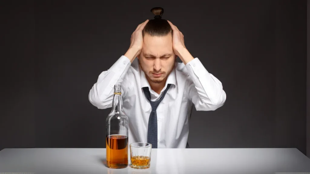 Young man in office attire holding his head in pain while sitting beside a bottle and glass of whiskey, depicting morning hangover symptoms after a party – ideal for articles on anti hangover solutions, morning recovery drinks, or after-party medicine in India.